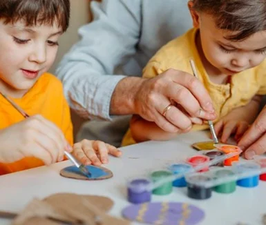 Zwei Kinder und eine erwachsene Person bemalen mit Pinseln Ostereier aus Pappe