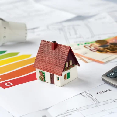 Ein Modellhaus liegt auf einem Energieausweis, Bauplänen und Geldscheinen.