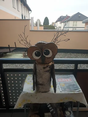 Eine aus Holz gebastelte Eule mit großen Augen steht auf einem Balkontisch.
